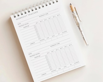 May include: A white spiral-bound workout tracker notebook with the words "WORKOUT TRACKER" printed at the top. A silver pen with a marble-like design rests beside the notebook. The notebook has sections for date, focus, exercise, and workout time.