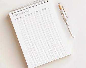 May include: A white spiral-bound medication log notebook with the words "MEDICATION LOG" printed at the top. A silver pen with a marbled design and wooden top rests beside the notebook. The notebook has columns for date, time, medication, dose, and notes.