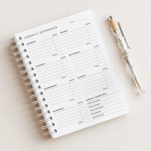 Puede incluir: Un cuaderno blanco con espiral con las palabras "WEEKLY EXPENSES" impresas en la parte superior. El cuaderno tiene secciones para cada día de la semana y una descripción general semanal. Un bolígrafo con un cuerpo transparente y detalles plateados descansa junto al cuaderno.