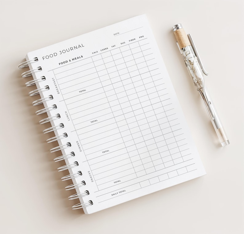 May include: A white spiral-bound food journal with the words "FOOD JOURNAL" printed at the top. The journal has a grid layout for tracking meals and nutrients. A clear pen with a decorative top rests beside the journal.