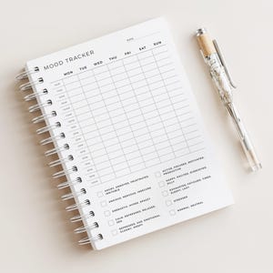 May include: A white spiral-bound mood tracker notebook with the title "MOOD TRACKER" and a grid for daily entries. A clear pen with a marbled design and a tan grip rests beside the notebook. The notebook includes checkboxes for mood descriptions.