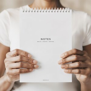 May include: A white spiral-bound notebook with the word "NOTES" printed in black, along with the text "WRITE. CREATE. INSPIRE." The notebook is held by a person wearing a white shirt. The brand name "MONVELLE" is printed at the bottom.