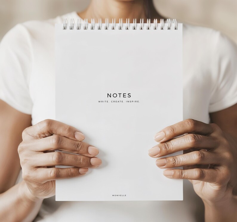 May include: A white spiral-bound notebook with the word "NOTES" printed in black, along with the text "WRITE. CREATE. INSPIRE." The notebook is held by a person wearing a white shirt. The brand name "MONVELLE" is printed at the bottom.