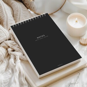 May include: A black spiral-bound notebook with the word "NOTES" and the text "WRITE CREATE INSPIRE" printed in white. The notebook is on top of a white book, next to a lit candle in a white holder. The word "MONVELLE" is printed at the bottom of the notebook.