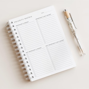 Può includere: Un taccuino bianco a spirale con la scritta "PRIORITY MATRIX". Il taccuino è aperto su una pagina con un layout a griglia e le intestazioni "IMPORTANT & URGENT", "IMPORTANT & NOT URGENT", "NOT IMPORTANT & URGENT" e "NOT IMPORTANT & NOT URGENT". Una penna con fusto trasparente e dettagli argentati è appoggiata accanto al taccuino.