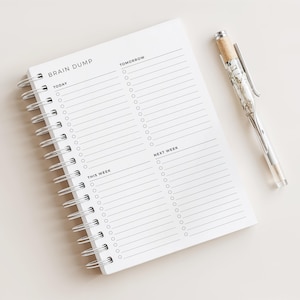 May include: A white spiral-bound notebook labelled "BRAIN DUMP" with sections for "TODAY", "TOMORROW", "THIS WEEK", and "NEXT WEEK". A clear pen with a silver clip and cork detail lies next to the notebook, ideal for planning and organisation.
