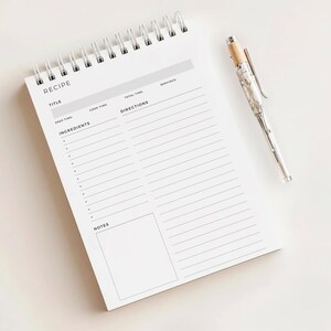 May include: A white recipe notebook with a spiral binding and a pen. The notebook has sections for title, ingredients, and directions. The pen is silver with a marbled design and a wooden top. The word "RECIPE" is printed at the top of the page.