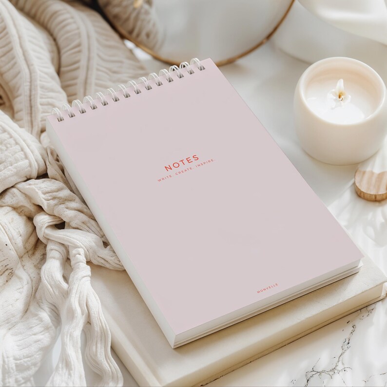 May include: A light pink spiral-bound notebook with the word "NOTES" printed in red. The text "WRITE. CREATE. INSPIRE." is below the word "NOTES". A white candle and a small wooden heart are in the background.