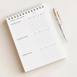 May include: A white spiral-bound habit tracker notebook with the words "HABIT TRACKER" at the top. The notebook has sections for morning, afternoon, and evening habits. A silver pen with a marble design rests beside the notebook.