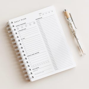 May include: A white spiral-bound daily planner with black text and lines. The page includes sections for a mood tracker, three priorities, a to-do list, a schedule from 5:30 am to 10:00 pm, meals, water intake, wellness, and gratitude. A clear pen with a tan grip sits beside it.