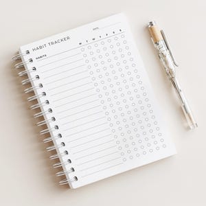 以下が含まれることがあります： 「HABIT TRACKER」と「HABITS」の文字が表紙に印刷された、白いリング綴じの習慣トラッカーノート。銀色のクリップと装飾的な上部が付いた透明なペンがノートの横に置かれています。ノートには、毎日の習慣を追跡するためのグリッドがあります。