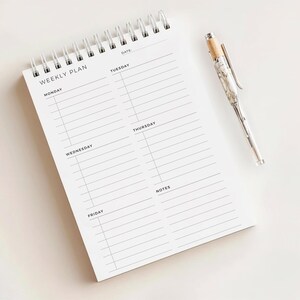 May include: A white weekly planner notepad with a silver spiral binding and a matching pen. The notepad is labelled "WEEKLY PLAN" and has sections for each day of the week, plus a notes section. The pen has a wood-tone top and a marble-like design.