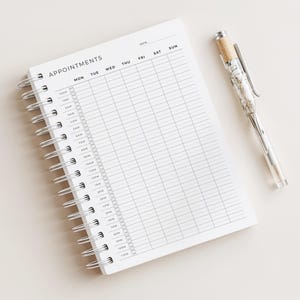 以下が含まれることがあります： 「APPOINTMENTS」の文字が上部に書かれた、白いリング綴じの予定ノート。銀色のクリップとコルクのような上部が付いた透明なペンがノートの横に置かれています。ノートはスケジュール用のグリッドレイアウトになっています。