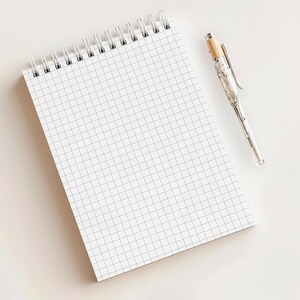 May include: A white grid-lined notepad with a silver spiral binding and a silver pen with a wood-tone top. The pen has a marble-like design. The notepad and pen are on a white surface.