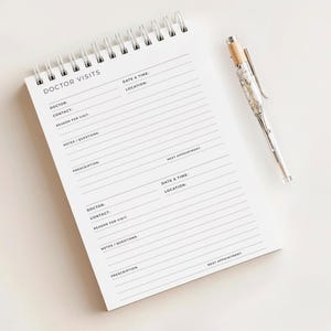 Könnte beinhalten: Ein weißer Spiralblock mit der Aufschrift "DOCTOR VISITS" oben. Er hat linierte Abschnitte zur Aufzeichnung von Arztinformationen, Termindetails und Notizen. Ein silberner Stift mit marmorähnlichem Design liegt daneben.