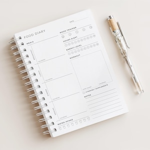 Può includere: Un diario alimentare bianco a spirale con la scritta "FOOD DIARY" in alto. Il diario include sezioni per i pasti, il monitoraggio dell'umore e i registri degli allenamenti. Una penna con un fusto trasparente e dettagli argentati è appoggiata accanto al diario.