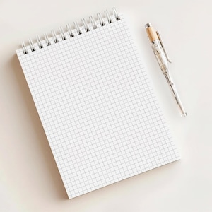 Könnte beinhalten: Ein weißes, kariertes Notizbuch mit silberner Spiralbindung und einem silbernen Stift mit Holzoptik. Der Stift hat ein marmorähnliches Design. Das Notizbuch und der Stift liegen auf einer weißen Oberfläche.