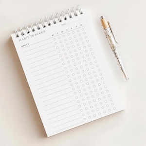 May include: A white spiral-bound habit tracker notebook with the words "HABIT TRACKER" and "HABITS" printed in black. A silver pen with a wood-tone top rests beside the notebook. The notebook has circles for tracking daily habits.