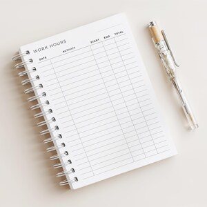 Könnte beinhalten: Ein weißes Spiralnotizbuch mit der Aufschrift "WORK HOURS". Es hat Spalten für Datum, Aktivität, Start, Ende und Gesamt. Ein durchsichtiger Stift mit silbernem Clip und beigefarbenem Oberteil liegt daneben.