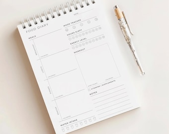 May include: A white spiral-bound food diary with the words "FOOD DIARY" at the top. The diary includes sections for meals, mood tracking, and workout logs. A silver pen with a wood-tone top rests beside the diary.