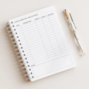 Könnte beinhalten: Ein weißes, spiralgebundenes Scheckbuchregister mit dem Aufdruck "CHECKBOOK REGISTER" oben. Das Register hat Spalten für Datum, Beschreibung, Gutschrift, Lastschrift und Saldo. Ein dekorativer Stift mit transparentem Schaft und silbernen Akzenten liegt neben dem Register.