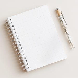 May include: A white spiral-bound notebook with dot grid pages and a silver pen with a clear barrel and cork-like top. The pen is lying on the right side of the notebook. The notebook and pen are on a light beige surface.