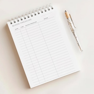 May include: A white spiral-bound notepad with the words "BLOOD PRESSURE" printed at the top. The notepad has columns for date, time, and blood pressure readings. A pen with a silver barrel and a wood-tone top rests beside the notepad.