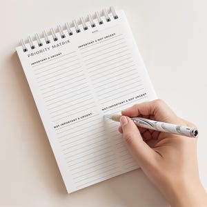 May include: A white spiral-bound notepad with the words "PRIORITY MATRIX" printed at the top. The notepad is open, with lines for writing. A silver and white pen is held over the notepad, ready to write.