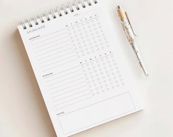 May include: A white spiral-bound skincare tracker notebook with the word "SKINCARE" at the top. The notebook has sections for morning and evening routines, with circles for tracking daily progress. A silver pen with a wood-tone top rests beside it.