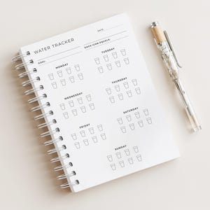 Op de afbeelding: Een wit spiraalgebonden water tracker notitieboek met de woorden "WATER TRACKER" bovenaan gedrukt. Het notitieboek heeft secties voor elke dag van de week, met ruimtes om de waterinname bij te houden. Een pen met een marmeren ontwerp ligt naast het notitieboek.