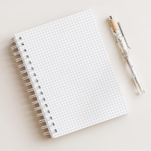 May include: A white spiral-bound notebook with a grid pattern and a silver pen with a clear barrel and a cork top. The pen is lying on a white surface next to the notebook.