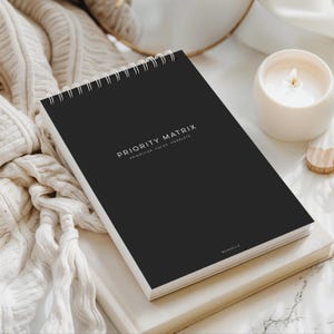 May include: A black spiral-bound notebook with the words "PRIORITY MATRIX" printed in white. The notebook is open, and the text "PRIORITIZE. FOCUS. COMPLETE" is below the title. A white candle and a beige blanket are in the background.