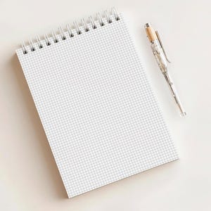 May include: A white, spiral-bound notebook with a grid pattern is positioned beside a silver pen with a marbled design and a wooden top. The notebook is open, revealing the grid. The pen is angled towards the notebook.