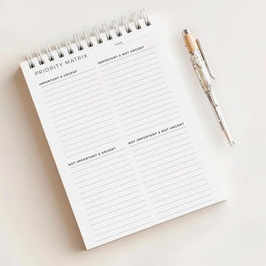 Può includere: Un taccuino bianco a spirale con la scritta "PRIORITY MATRIX" stampata in nero. Il taccuino è aperto, rivelando pagine a righe per organizzare le attività. Una penna d'argento con la parte superiore in legno è appoggiata accanto al taccuino.