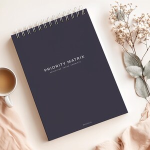 May include: A dark navy blue spiral-bound notebook with the words "PRIORITY MATRIX" printed in white. The notebook is open, and a cup of tea and decorative flowers are next to it. The notebook's text also includes "PRIORITIZE. FOCUS. COMPLETE."