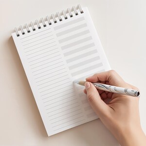 May include: A white spiral-bound notepad with lined and columned pages. A silver and white pen is held by a hand, poised to write on the notepad. The notepad is open and ready for use.