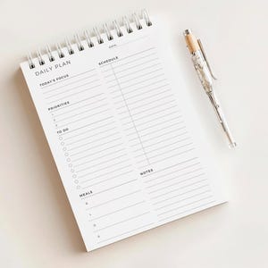 May include: A white spiral-bound daily plan notepad with the words "DAILY PLAN" printed at the top. The notepad includes sections for "Today's Focus," "Priorities," "To Do," "Schedule," "Meals," and "Notes." A silver pen with a wood-tone top rests on the right side.
