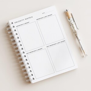 Op de afbeelding: Een wit spiraalgebonden notitieboek met de tekst "PRIORITY MATRIX". Het notitieboek is open op een pagina met vier secties met de labels "Important & Urgent", "Important & Not Urgent", "Not Important & Urgent" en "Not Important & Not Urgent". Een doorzichtige pen met een decoratief ontwerp ligt naast het notitieboek.