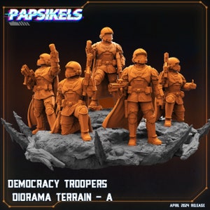 Op de afbeelding: Een groep oranje Democracy Troopers-beeldjes, geposeerd op een grijs diorama-terrein. De figuren dragen helmen en harnassen en houden wapens vast. De afbeelding bevat de tekst "Papsikels", "Democracy Troopers", "Diorama Terrain - A" en "April 2024 Release".
