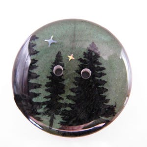 May include: A round button with a forest scene. The button features two dark green pine trees, silver and gold star accents, and two button holes. The background is a muted green.