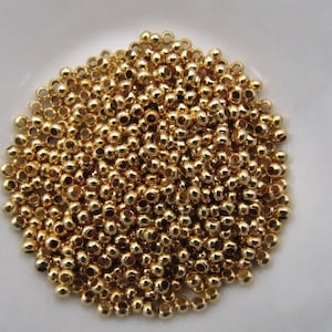 May include: A pile of small, shiny, gold-colored beads. The beads are round with a small hole through the center, and they are scattered across a white surface. These beads are likely used for jewelry making or other crafts.