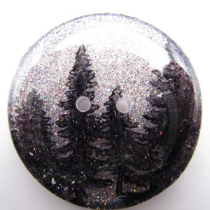 May include: A round button with a forest scene. The button has a clear, glittery top with a black forest silhouette design. Two holes are visible for sewing. The button is set against a white background.