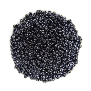 May include: A circular pile of small, round, dark metallic grey seed beads. Each individual bead features a smooth, reflective surface with a central hole, creating a uniform appearance across the entire collection of loose craft components.