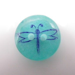 Puede incluir: Un botón redondo turquesa con un diseño de libélula azul oscuro. La libélula está dibujada a mano con líneas finas y tiene dos pequeños ojales blancos. Suministro de artesanía.
