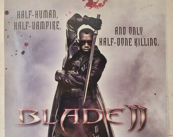May include: A movie poster for the film Blade II, featuring a man in a black leather coat and sunglasses holding a sword. The text on the poster reads "HALF-HUMAN, HALF-VAMPIRE, AND ONLY HALF-DONE KILLING. BLADE II"