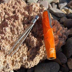 May include: A close-up of a silver and orange pen with a swirl pattern. The pen is lying on a textured, light brown surface.