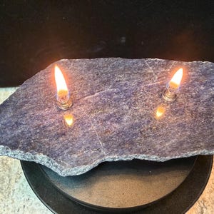 May include: A natural, blue-grey stone with two lit flames. The stone sits on a black, circular base. The flames are reflected in the stone's surface, creating a warm, inviting ambiance. This item is a decorative candle holder.