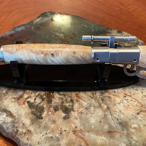 May include: A unique pen resembling a rifle, featuring a light-colored, patterned wooden barrel and a silver-colored metal mechanism. The pen includes a scope and is displayed on a black stand, set against a stone surface.