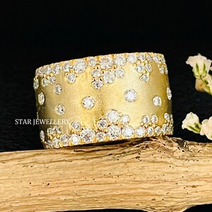 Può includere: Un anello largo, color oro, con una superficie strutturata, incrostato con numerose gemme trasparenti. L'anello è posto su uno sfondo scuro, con un rametto di fiori bianchi a destra. L'anello è contrassegnato dal testo "STAR JEWELLERY".