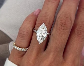 May include: A marquise-cut diamond engagement ring with a silver band. A smaller gold band with small diamonds is also visible. The hand has pink nail polish.